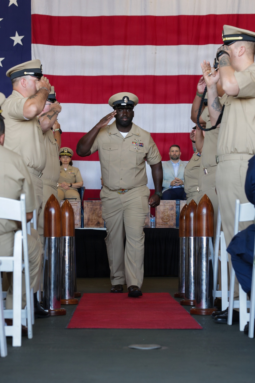 A U.S. Navy Chief Petty Officer in khaki service uniform walks down a red carpet aisle while rendering a salute during what appears to be a Chief Petty Officer pinning or retirement ceremony. He is flanked by rows of fellow sailors in khaki uniforms who are applauding, with decorative shell casing pedestals lining the aisle. A large American flag serves as the backdrop for the formal military ceremony.