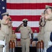 A U.S. Navy Chief Petty Officer in khaki service uniform walks down a red carpet aisle while rendering a salute during what appears to be a Chief Petty Officer pinning or retirement ceremony. He is flanked by rows of fellow sailors in khaki uniforms who are applauding, with decorative shell casing pedestals lining the aisle. A large American flag serves as the backdrop for the formal military ceremony.