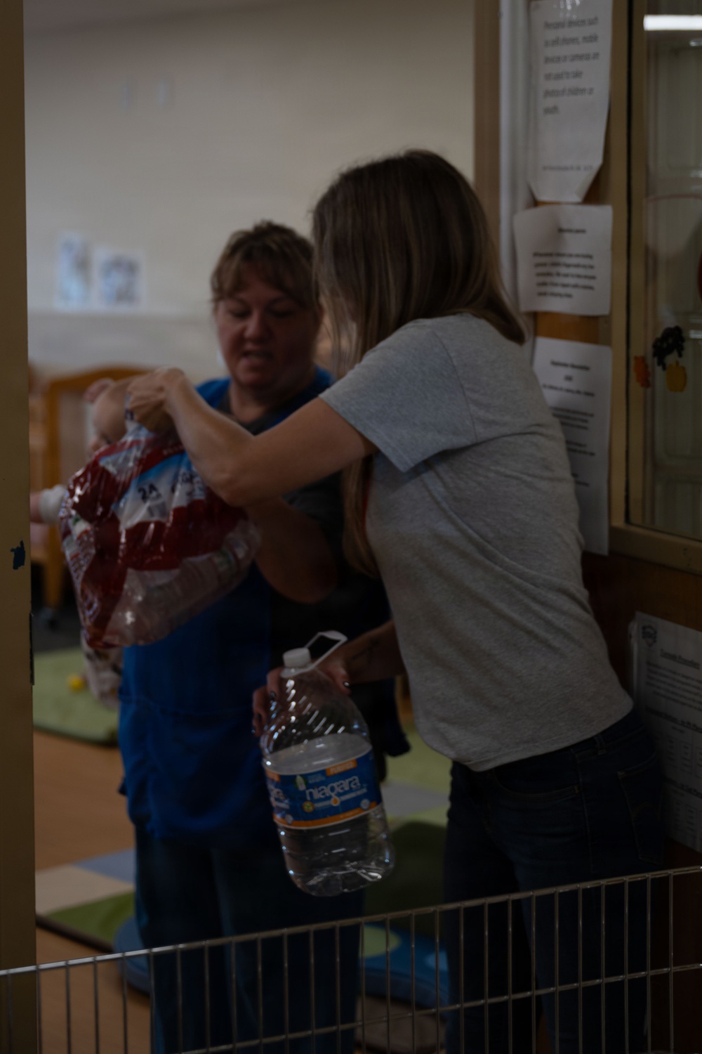 Two women are interacting in what appears to be a childcare or classroom setting. One woman in a blue top is handing or receiving a red packaged item, while the other woman in a gray t-shirt is holding a Niagara brand water bottle. Several posted notices or papers are visible on a wooden bulletin board to the right.