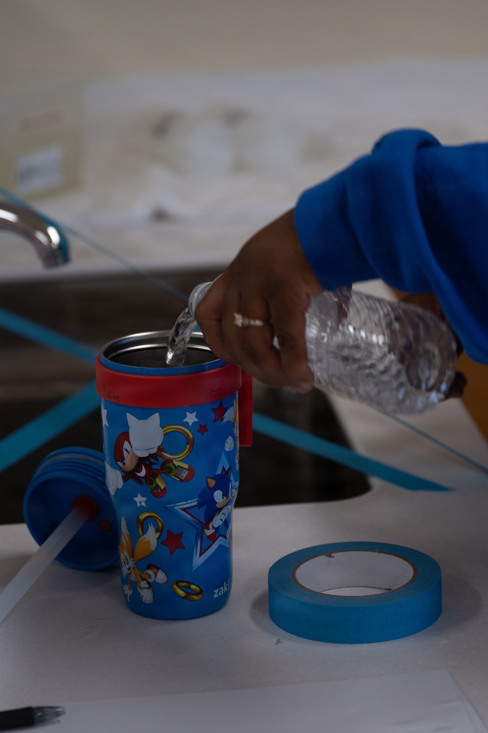 A person wearing a blue long-sleeve shirt is pouring water from a clear plastic bottle into a Zak! brand children's insulated tumbler featuring Sonic the Hedgehog characters. The scene takes place near a kitchen or bathroom sink, with blue painter's tape and the tumbler's lid and straw visible on the white surface. The cup's lid has been removed and set aside to allow filling.