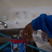A person wearing a blue long-sleeve shirt is pouring water from a clear plastic bottle into a Zak! brand children's insulated tumbler featuring Sonic the Hedgehog characters. The scene takes place near a kitchen or bathroom sink, with blue painter's tape and the tumbler's lid and straw visible on the white surface. The cup's lid has been removed and set aside to allow filling.