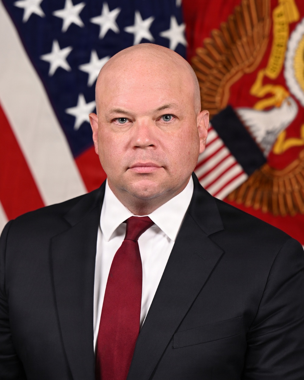 A formal official portrait of a bald middle-aged man wearing a dark suit with a white dress shirt and dark red/burgundy tie. He is posed in front of an American flag and what appears to be a red military or departmental seal flag. The setting and composition are consistent with a U.S. government or military official portrait.