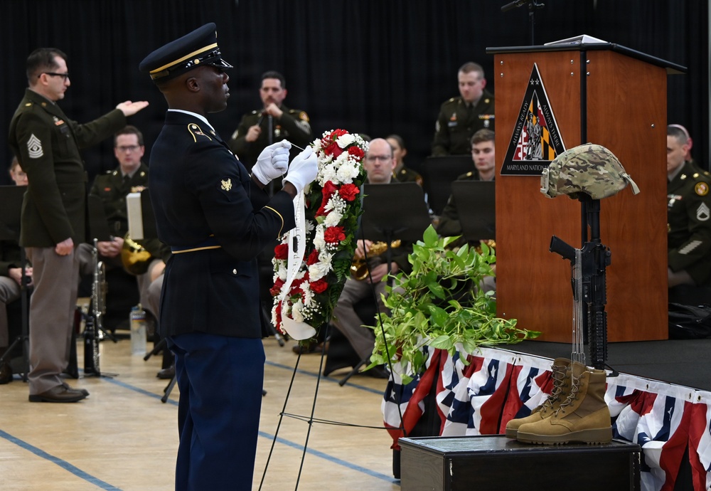 A military memorial ceremony is taking place in what appears to be a gymnasium or indoor venue. A soldier in dress uniform is presenting a red, white, and green floral wreath near a battlefield cross display consisting of boots, a rifle, and a combat helmet. A military band and other uniformed personnel are visible in the background near a podium bearing a Maryland National Guard insignia.