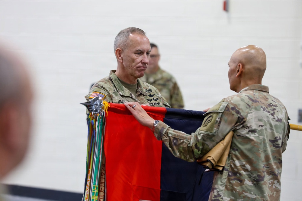 Two U.S. Army soldiers in OCP (Operational Camouflage Pattern) uniforms are participating in a military colors ceremony, handling a folded guidon or unit flag with streamers attached. The soldier on the left, whose name tape reads 'JAMES,' appears to be a senior officer receiving or passing the colors. This type of ceremony typically marks a change of command or unit activation/inactivation.