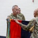 Two U.S. Army soldiers in OCP (Operational Camouflage Pattern) uniforms are participating in a military colors ceremony, handling a folded guidon or unit flag with streamers attached. The soldier on the left, whose name tape reads 'JAMES,' appears to be a senior officer receiving or passing the colors. This type of ceremony typically marks a change of command or unit activation/inactivation.