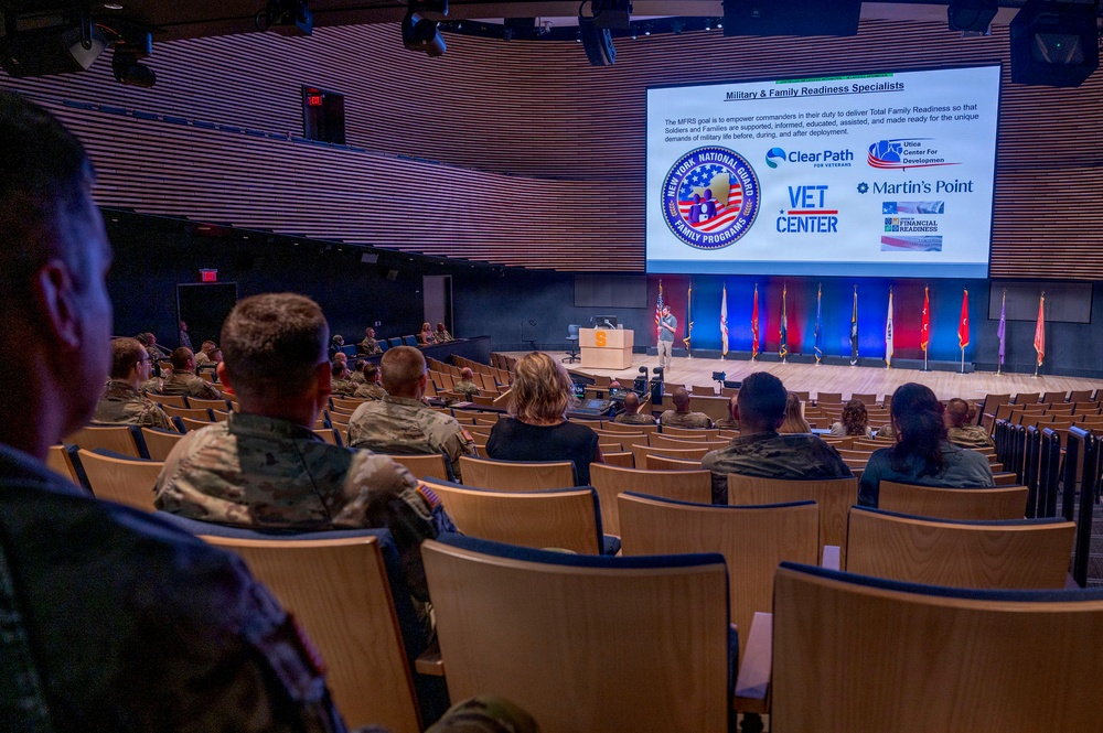 A military briefing or training event is taking place in a modern auditorium, with an audience composed primarily of uniformed military personnel and some civilians. A presenter stands at a podium on stage in front of a large projection screen displaying a slide titled 'Military &amp; Family Readiness Specialists' featuring the New York National Guard Family Programs logo and partner organization logos. The seating is partially filled with attendees spread across the tiered auditorium seating.