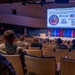 A military briefing or training event is taking place in a modern auditorium, with an audience composed primarily of uniformed military personnel and some civilians. A presenter stands at a podium on stage in front of a large projection screen displaying a slide titled 'Military &amp; Family Readiness Specialists' featuring the New York National Guard Family Programs logo and partner organization logos. The seating is partially filled with attendees spread across the tiered auditorium seating.
