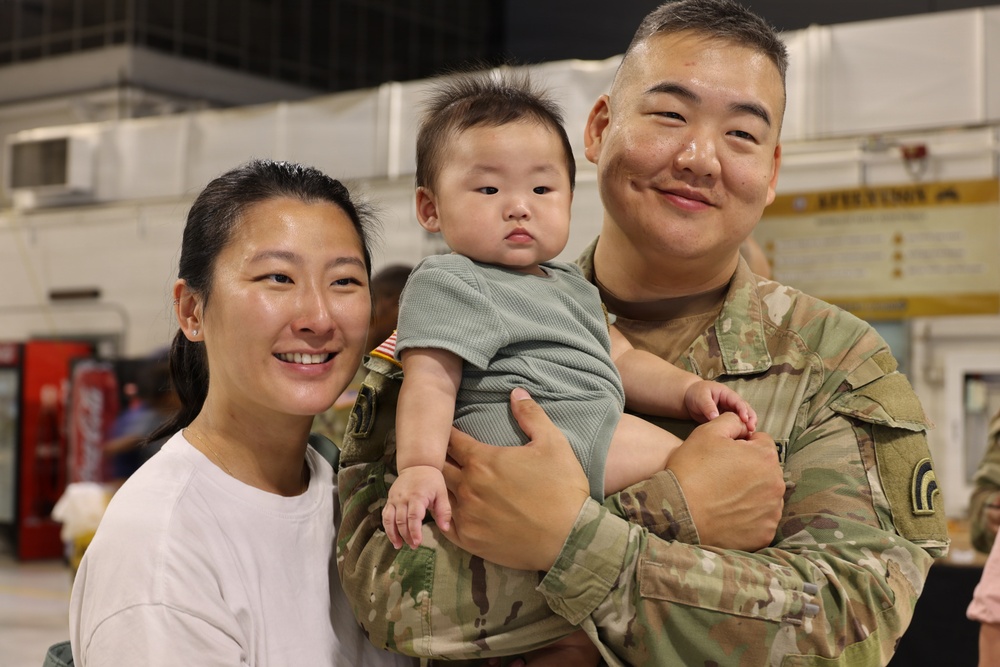 A military service member in U.S. Army OCP (Operational Camouflage Pattern) uniform holds a young infant while standing next to a woman in a white shirt. The setting appears to be an indoor military facility, likely a hangar or gymnasium, suggesting a homecoming or deployment event. All three subjects are facing the camera in what appears to be a family portrait moment.