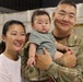 A military service member in U.S. Army OCP (Operational Camouflage Pattern) uniform holds a young infant while standing next to a woman in a white shirt. The setting appears to be an indoor military facility, likely a hangar or gymnasium, suggesting a homecoming or deployment event. All three subjects are facing the camera in what appears to be a family portrait moment.