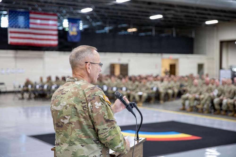 A U.S. Army soldier in OCP (Operational Camouflage Pattern) uniform addresses a large formation of seated soldiers in what appears to be a military armory or gymnasium. The speaker stands at a wooden podium with a microphone, facing an audience of dozens of uniformed personnel. American and unit flags are visible hanging in the background.