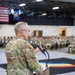 A U.S. Army soldier in OCP (Operational Camouflage Pattern) uniform addresses a large formation of seated soldiers in what appears to be a military armory or gymnasium. The speaker stands at a wooden podium with a microphone, facing an audience of dozens of uniformed personnel. American and unit flags are visible hanging in the background.