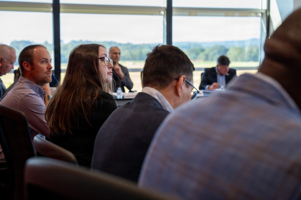 A group of people are seated at tables in a modern conference or meeting room with large floor-to-ceiling windows overlooking a green outdoor landscape. Attendees appear to be listening attentively to a presentation or speaker outside the frame. The setting suggests a formal business or professional gathering.