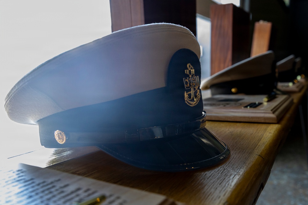A formal naval dress cap with a white crown and dark navy band is displayed on a wooden surface. The cap features a gold anchor insignia on the front and a small gold button on the side, consistent with U.S. Navy Chief Petty Officer or officer dress uniform headgear. Additional caps and wooden display items are visible in the background.