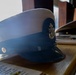 A formal naval dress cap with a white crown and dark navy band is displayed on a wooden surface. The cap features a gold anchor insignia on the front and a small gold button on the side, consistent with U.S. Navy Chief Petty Officer or officer dress uniform headgear. Additional caps and wooden display items are visible in the background.