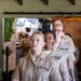 The image shows a group of U.S. Navy personnel in Service Khaki uniforms walking in formation through what appears to be a restaurant or event venue. At least three women are prominently visible in the foreground, each wearing ribbons, warfare devices, and other military insignia on their uniforms. Additional personnel in Navy Working Uniform (NWU) camouflage are visible in the background.