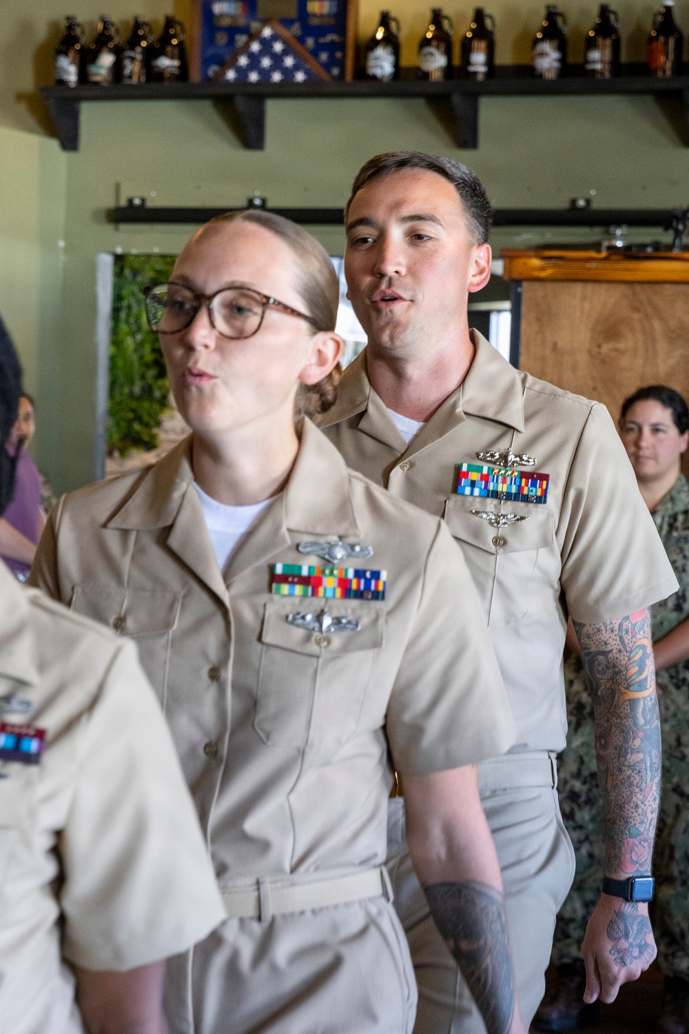 Two U.S. Navy personnel in Service Khaki uniforms (Service Uniform Alpha) are standing indoors, appearing to be speaking or reciting something simultaneously. Both wear multiple service ribbons, warfare qualification badges, and other insignia on their uniforms. The setting appears to be a casual venue, possibly a brewery or taproom, with bottle growlers and a folded American flag visible on shelving in the background.