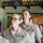 Two U.S. Navy personnel in Service Khaki uniforms (Service Uniform Alpha) are standing indoors, appearing to be speaking or reciting something simultaneously. Both wear multiple service ribbons, warfare qualification badges, and other insignia on their uniforms. The setting appears to be a casual venue, possibly a brewery or taproom, with bottle growlers and a folded American flag visible on shelving in the background.
