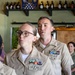 Two U.S. Navy personnel in Service Khaki uniforms (Service Uniform Alpha) are standing indoors, appearing to be speaking or reciting something simultaneously. Both wear multiple service ribbons, warfare qualification badges, and other insignia on their uniforms. The setting appears to be a casual venue, possibly a brewery or taproom, with bottle growlers and a folded American flag visible on shelving in the background.