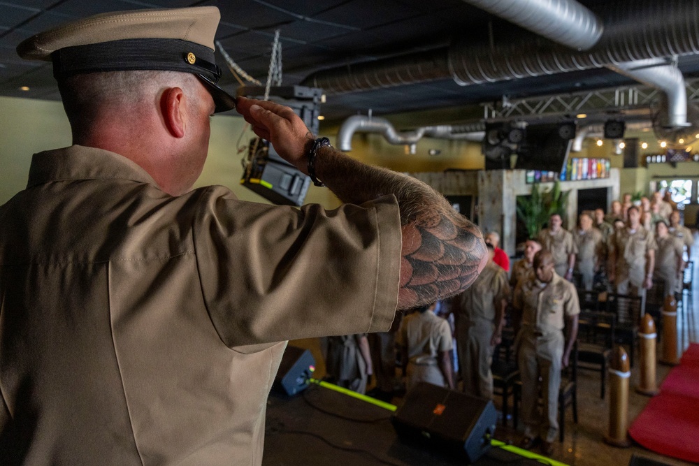 A U.S. Navy enlisted service member in khaki uniform is saluting from a stage or elevated position toward a formation of fellow sailors standing at attention in what appears to be a converted venue or event space. The individual in the foreground has a prominent tattoo sleeve on their right arm and is wearing a combination cover (white cap). This appears to be a military ceremony, possibly a promotion, retirement, or change of command event.