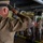 A U.S. Navy enlisted service member in khaki uniform is saluting from a stage or elevated position toward a formation of fellow sailors standing at attention in what appears to be a converted venue or event space. The individual in the foreground has a prominent tattoo sleeve on their right arm and is wearing a combination cover (white cap). This appears to be a military ceremony, possibly a promotion, retirement, or change of command event.