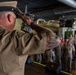 A U.S. Navy enlisted service member in khaki uniform is saluting from a stage or elevated position toward a formation of fellow sailors standing at attention in what appears to be a converted venue or event space. The individual in the foreground has a prominent tattoo sleeve on their right arm and is wearing a combination cover (white cap). This appears to be a military ceremony, possibly a promotion, retirement, or change of command event.