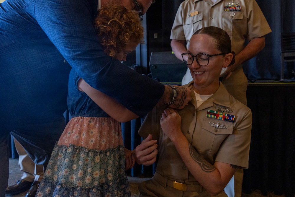 A U.S. Navy service member in tan/khaki uniform is kneeling or crouching while a child and an adult pin insignia or devices onto her collar or shoulder during what appears to be a military promotion or pinning ceremony. The sailor is smiling and wearing glasses, with visible ribbons, medals, and a warfare device on her uniform. A senior Navy officer in the background observes the ceremony.