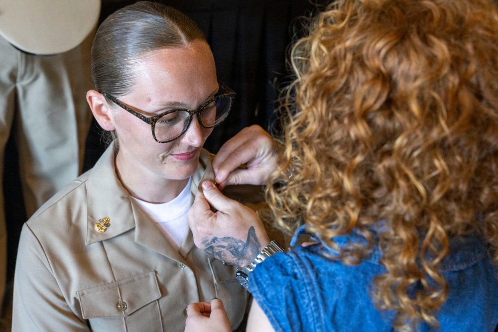 A woman in a tan/khaki military uniform is having an insignia or pin attached to her collar by another person with curly auburn hair and a visible tattoo on their hand. The uniformed individual is wearing glasses and has her hair pulled back neatly. This appears to be a military promotion or pinning ceremony.