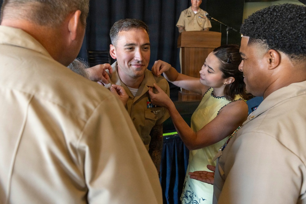A military promotion or pinning ceremony is taking place, with multiple individuals pinning new insignia or rank devices onto a uniformed service member's collar. The central figure, wearing tan/khaki U.S. Navy or Marine Corps service uniform with ribbons and a warfare device, is smiling as a young girl in a yellow dress and other uniformed personnel assist with the pinning. A senior officer stands at a podium in the background, suggesting a formal ceremony setting.