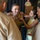 A military promotion or pinning ceremony is taking place, with multiple individuals pinning new insignia or rank devices onto a uniformed service member's collar. The central figure, wearing tan/khaki U.S. Navy or Marine Corps service uniform with ribbons and a warfare device, is smiling as a young girl in a yellow dress and other uniformed personnel assist with the pinning. A senior officer stands at a podium in the background, suggesting a formal ceremony setting.