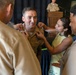 A military promotion or pinning ceremony is taking place, with multiple individuals pinning new insignia or rank devices onto a uniformed service member's collar. The central figure, wearing tan/khaki U.S. Navy or Marine Corps service uniform with ribbons and a warfare device, is smiling as a young girl in a yellow dress and other uniformed personnel assist with the pinning. A senior officer stands at a podium in the background, suggesting a formal ceremony setting.