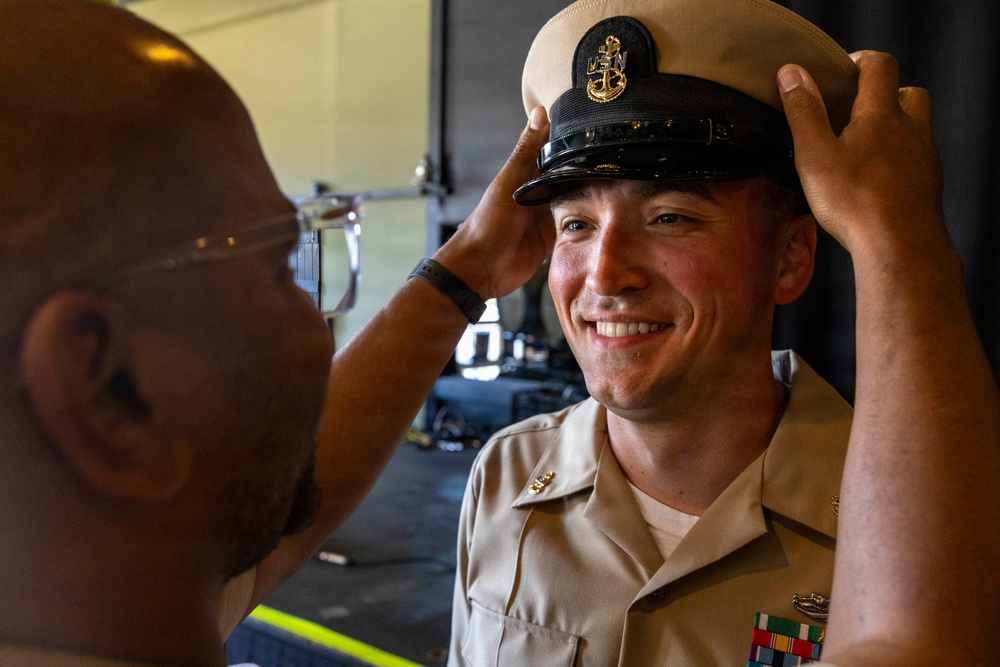 A U.S. Navy sailor is being fitted with a Chief Petty Officer combination cover (cap) by another individual, likely during a promotion or pinning ceremony. The smiling sailor is wearing a khaki service uniform with ribbons and collar devices. This appears to be a milestone military advancement event.