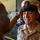 A U.S. Navy sailor is being fitted with a Chief Petty Officer combination cover (cap) by another individual, likely during a promotion or pinning ceremony. The smiling sailor is wearing a khaki service uniform with ribbons and collar devices. This appears to be a milestone military advancement event.