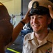A U.S. Navy sailor is being fitted with a Chief Petty Officer combination cover (cap) by another individual, likely during a promotion or pinning ceremony. The smiling sailor is wearing a khaki service uniform with ribbons and collar devices. This appears to be a milestone military advancement event.