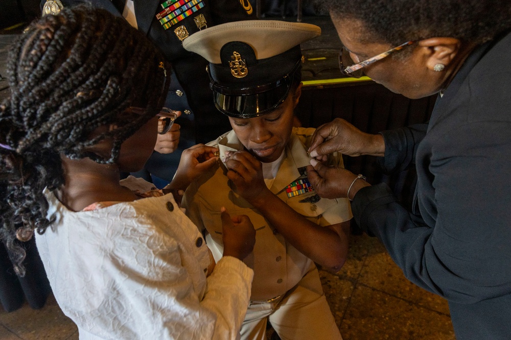 A military promotion or pinning ceremony is taking place, with a service member in U.S. Navy dress uniform being pinned by two individuals, likely family members. The central figure wears a white Navy service dress uniform with a combination cover (officer's cap), ribbons, and a badge. Two people — a young girl in a white dress and an older woman in dark clothing — are simultaneously pinning insignia onto the uniform's collar.