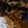 A military promotion or pinning ceremony is taking place, with a service member in U.S. Navy dress uniform being pinned by two individuals, likely family members. The central figure wears a white Navy service dress uniform with a combination cover (officer's cap), ribbons, and a badge. Two people — a young girl in a white dress and an older woman in dark clothing — are simultaneously pinning insignia onto the uniform's collar.