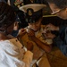 A military promotion or pinning ceremony is taking place, with a service member in U.S. Navy dress uniform being pinned by two individuals, likely family members. The central figure wears a white Navy service dress uniform with a combination cover (officer's cap), ribbons, and a badge. Two people — a young girl in a white dress and an older woman in dark clothing — are simultaneously pinning insignia onto the uniform's collar.