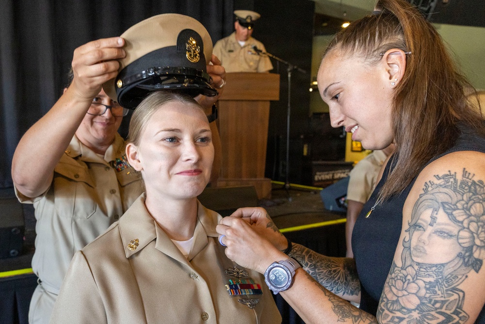 A military promotion or pinning ceremony is taking place, likely in the U.S. Navy. A service member in tan/khaki Navy uniform is being pinned with a new insignia by a civilian woman while another uniformed service member places a chief petty officer cover (hat) on her head. A speaker at a podium is visible in the background.