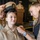 A military promotion or pinning ceremony is taking place, likely in the U.S. Navy. A service member in tan/khaki Navy uniform is being pinned with a new insignia by a civilian woman while another uniformed service member places a chief petty officer cover (hat) on her head. A speaker at a podium is visible in the background.