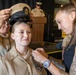 A military promotion or pinning ceremony is taking place, likely in the U.S. Navy. A service member in tan/khaki Navy uniform is being pinned with a new insignia by a civilian woman while another uniformed service member places a chief petty officer cover (hat) on her head. A speaker at a podium is visible in the background.