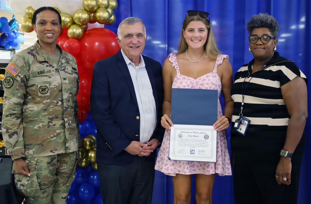 A scholarship presentation ceremony for military children is taking place in front of a blue backdrop with red, white, and gold balloon decorations. A young woman named Emily Royster is holding a 'Scholarships for Military Children' certificate, flanked by a U.S. Army soldier (name tag reads 'KING'), an older civilian man in a navy blazer, and a civilian woman in a striped top. The event appears to be held at a military installation commissary or community facility.