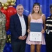 A scholarship presentation ceremony for military children is taking place in front of a blue backdrop with red, white, and gold balloon decorations. A young woman named Emily Royster is holding a 'Scholarships for Military Children' certificate, flanked by a U.S. Army soldier (name tag reads 'KING'), an older civilian man in a navy blazer, and a civilian woman in a striped top. The event appears to be held at a military installation commissary or community facility.