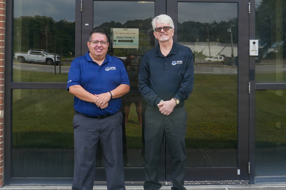 Two men wearing matching AFSC-branded polo shirts are standing in front of a glass-door entrance to a building. Both are posing for a photo with hands clasped in front of them. A sign on the door references a Visitor Center with a directional arrow.