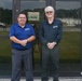 Two men wearing matching AFSC-branded polo shirts are standing in front of a glass-door entrance to a building. Both are posing for a photo with hands clasped in front of them. A sign on the door references a Visitor Center with a directional arrow.