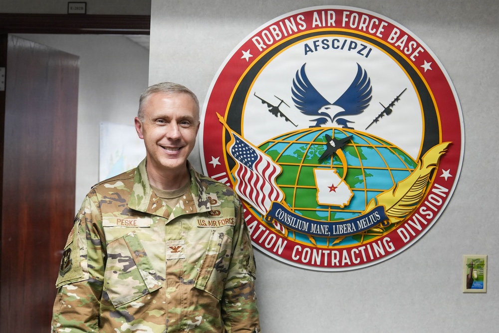 A U.S. Air Force officer in OCP (Operational Camouflage Pattern) uniform stands smiling beside an official unit emblem at Robins Air Force Base. The insignia identifies the location as Robins AFB and references the Installation Contracting Division under AFSC/PZI. The officer's name tape reads 'PESKE' and rank insignia (colonel eagle) is visible on the shoulder.