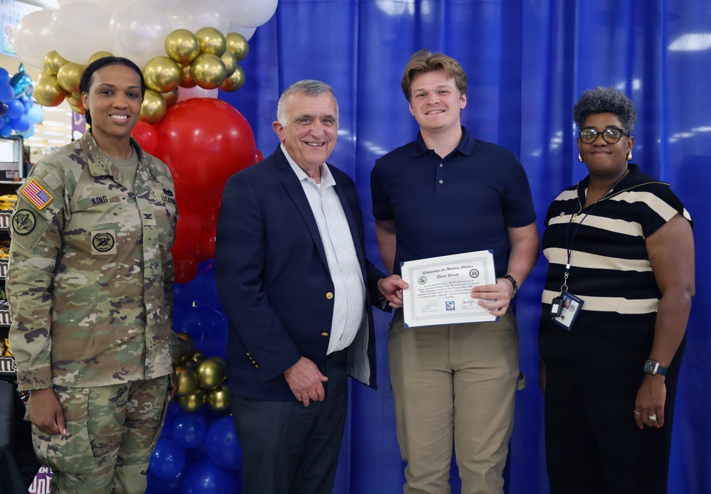 Four individuals pose for a formal photo at what appears to be a military scholarship award ceremony. A young man is holding a certificate that reads 'Scholarships for Military Children' while standing alongside a U.S. Army soldier in camouflage uniform with the name tape 'KING', an older civilian man in a navy blazer, and a civilian woman wearing a lanyard with an ID badge. The backdrop features red, white, blue, and gold balloons arranged decoratively against a blue curtain.