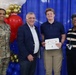 Four individuals pose for a formal photo at what appears to be a military scholarship award ceremony. A young man is holding a certificate that reads 'Scholarships for Military Children' while standing alongside a U.S. Army soldier in camouflage uniform with the name tape 'KING', an older civilian man in a navy blazer, and a civilian woman wearing a lanyard with an ID badge. The backdrop features red, white, blue, and gold balloons arranged decoratively against a blue curtain.