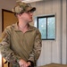A U.S. military service member in MultiCam (OCP) uniform stands indoors next to wooden ammunition crates. He is wearing tactical hearing protection mounted on a multicam cap, glasses, and a combat shirt, and appears to be holding a small device while looking upward. The setting appears to be a range facility or supply room.