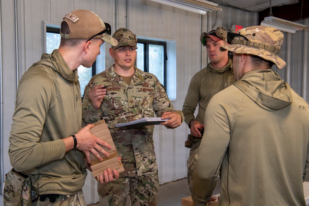 Four U.S. Air Force personnel in OCP (Operational Camouflage Pattern) uniforms are gathered inside a metal building, appearing to conduct a briefing or review session. The central figure, wearing a full OCP uniform with rank insignia and a name tape partially readable as 'BERNALD' or similar, is holding documents and appears to be speaking to the group. The others are in various combinations of OCP gear and olive drab athletic wear.