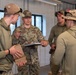 Four U.S. Air Force personnel in OCP (Operational Camouflage Pattern) uniforms are gathered inside a metal building, appearing to conduct a briefing or review session. The central figure, wearing a full OCP uniform with rank insignia and a name tape partially readable as 'BERNALD' or similar, is holding documents and appears to be speaking to the group. The others are in various combinations of OCP gear and olive drab athletic wear.