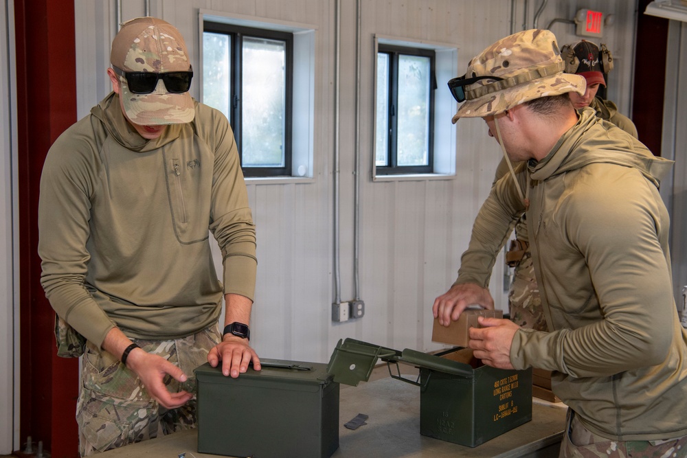 Two military personnel in multicam camouflage uniforms are working with olive drab ammunition cans on a table inside a building. A third individual is partially visible in the background. The setting appears to be a military facility or range building based on the environment and equipment present.